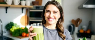 A woman smiling confidently while holding a healthy green smoothie in a bright, sunlit kitchen, surrounded by fresh vegetables and fruits, conveying wellness and vitality